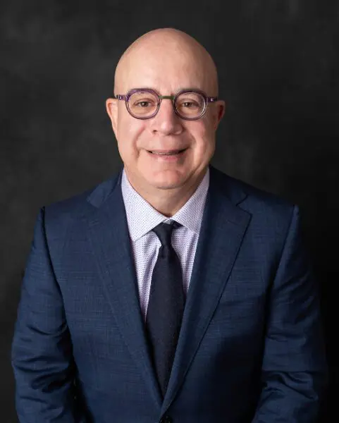 Portrait of a smiling man in a suit and glasses against a dark background.